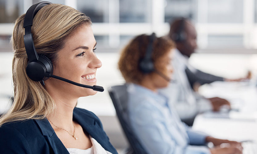 Three workers sitting in a row of desks in an office. Tey are all wearing phone headsets and looking at computer monitors.