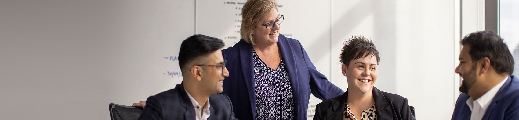 A group of people wearing suits. They are sitting around an office table, with one person standing.