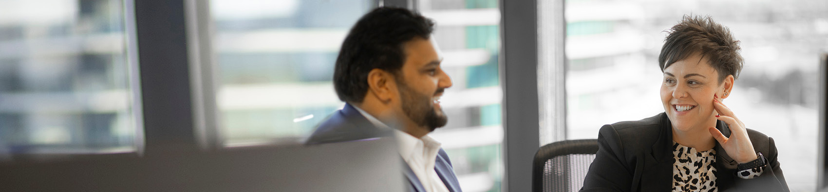 Two people wearing suits and sitting in an office by the window. They are both smiling.