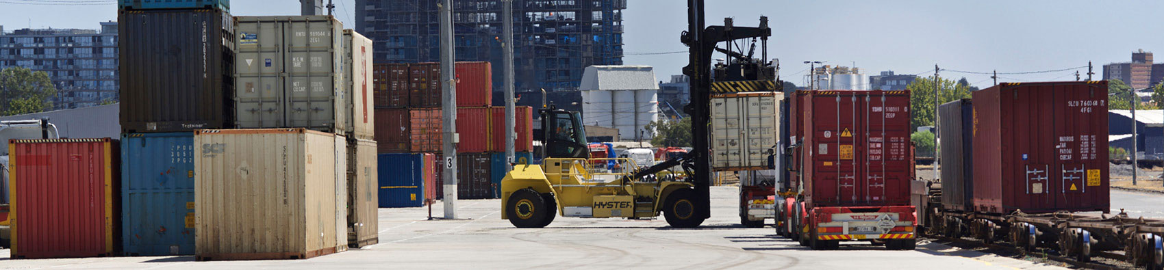 Dynon terminal with a yellow container handler that lifts and stacks shipping containers. The vehicle is surrounded by about 12 shipping containers.