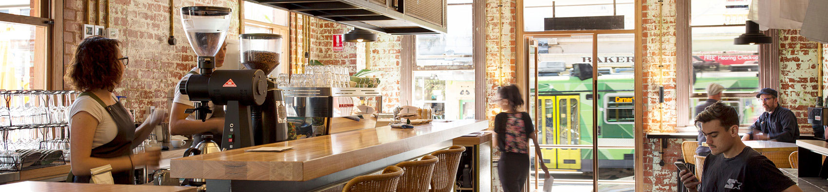 Inside a cafe that has an industrial look with exposed bricks. There are two workers behind the counter, and some customers sitting at tables.