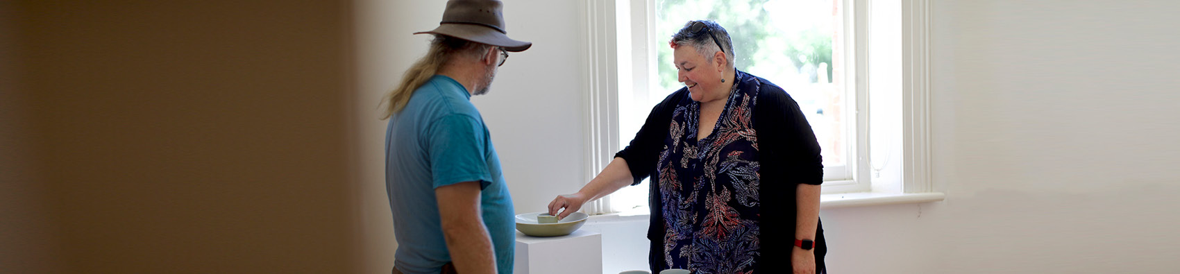 Two people in a community arts gallery. One person is touching an artwork on a wooden pedestal. 