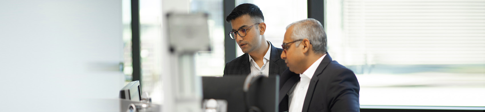 Two people wearing suits and standing in an office and looking at a computer. There are large windows in the background.