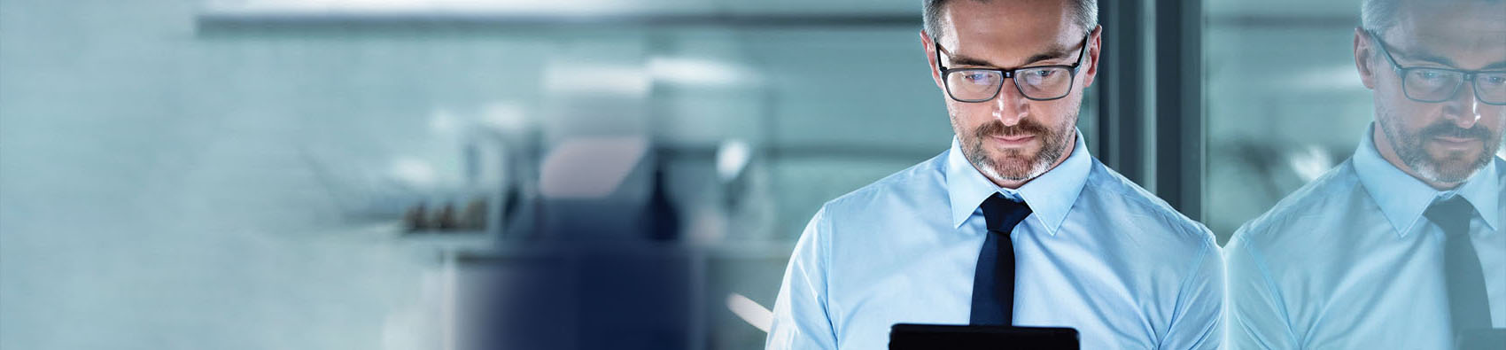 A person wearing business attire leaning against a glass wall in an office, and reading a tablet.