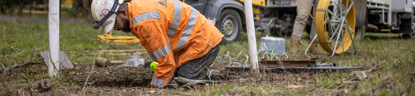 A worker wearing a hi vis safety jacket and white helmet. He is working in a hole in the ground. There is grass around him.