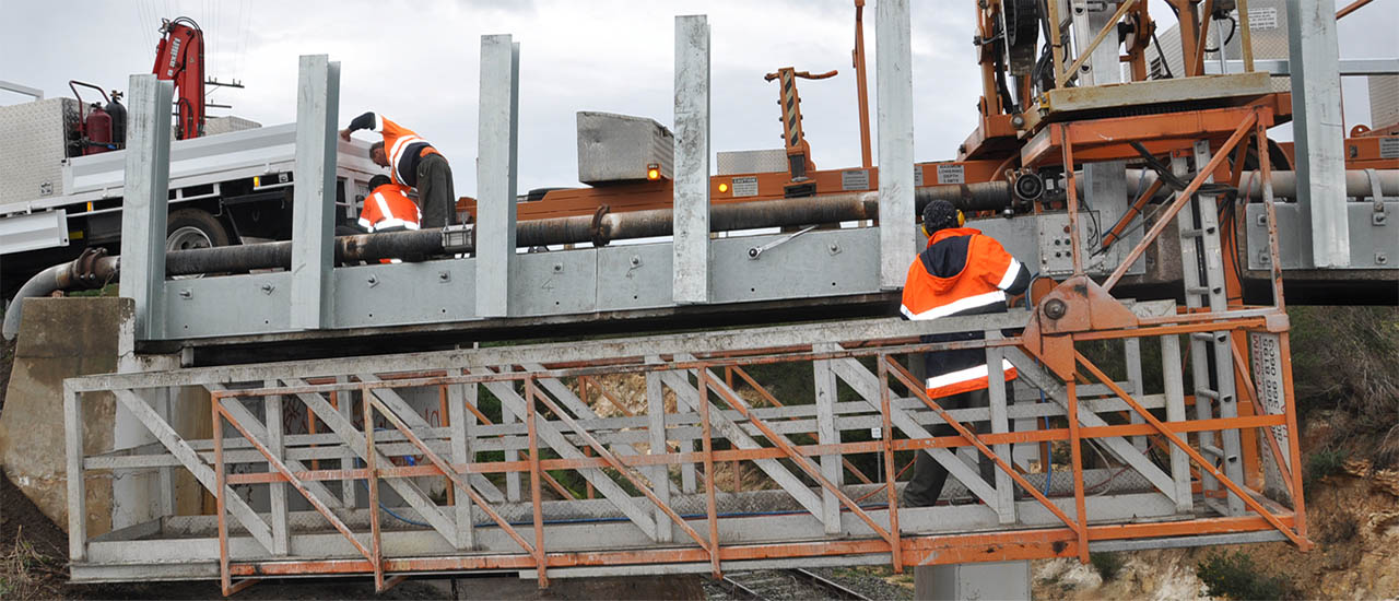 Two workers wearing hi vis safety jackets working on a metail rail bridge. A third worker is in the foreground and standing on an under bridge boom and working lower on the bridge.