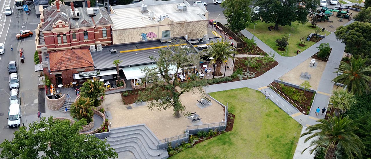 An aerial view of the Elsternwick Station precinct. A restaurant bar with tables and chairs outside is in the background. There is a grass section at the front and the grass area is surrounded by trees.