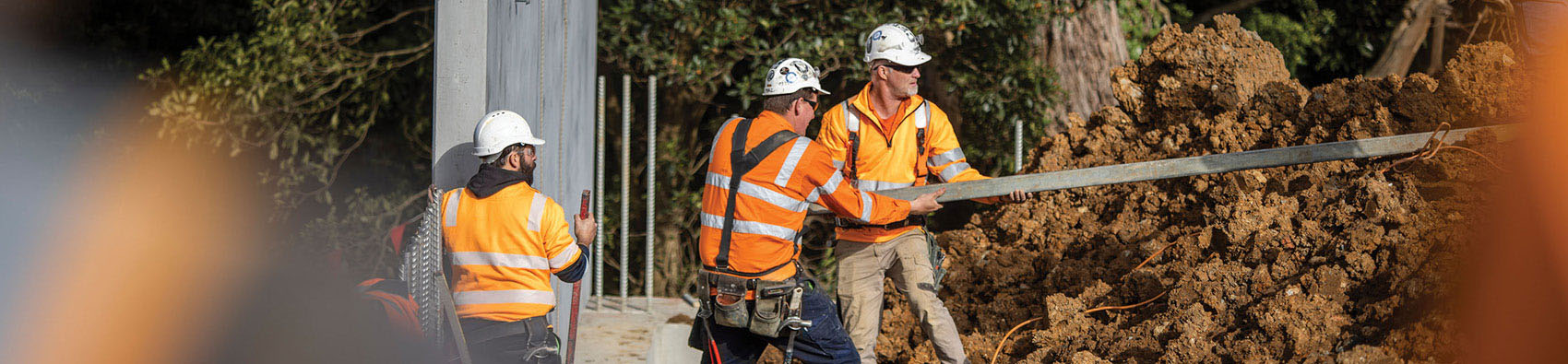 Three workers wearing orange hi vis jackets and safety helmets. They are working outdoors and there is a concrete beam and a pile of dirt. Two workers are lifting a wooden beam.