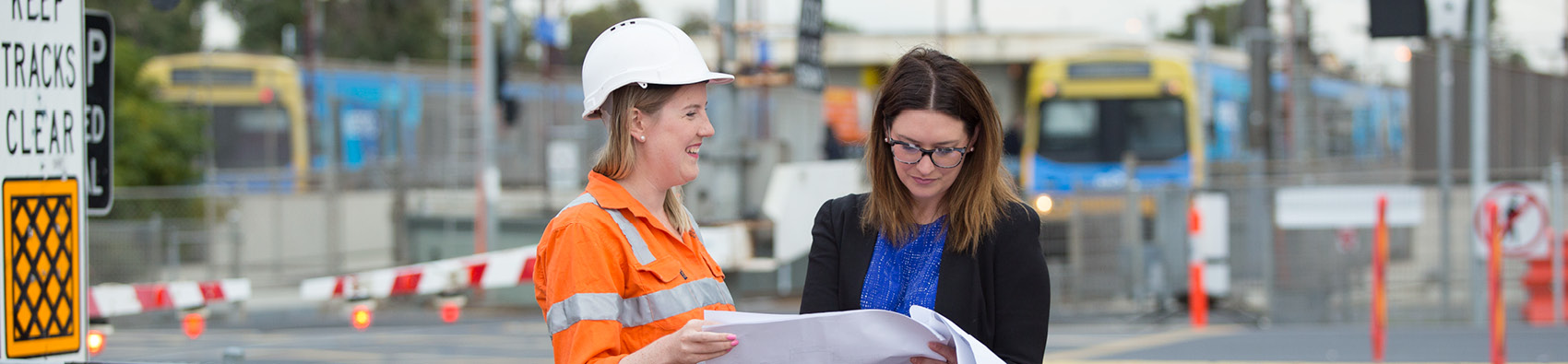 Two workers holding documents outside a train level crossing. The boomgate is down and a train is in the background.