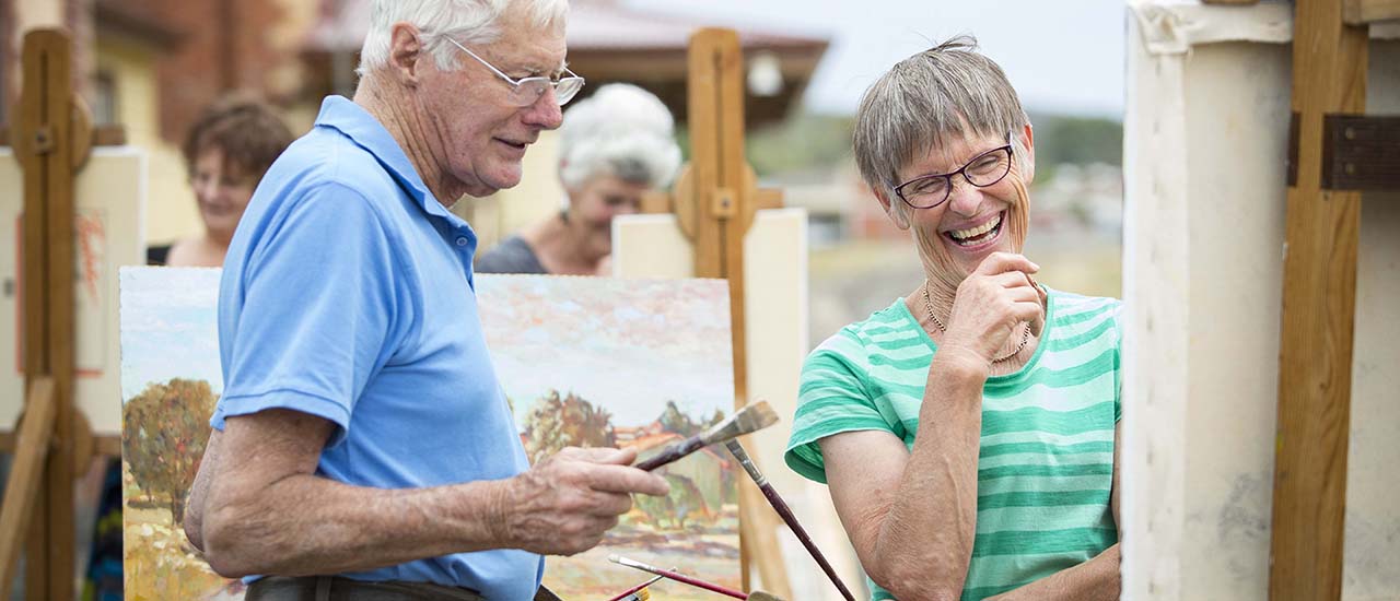 To people admire a painting that is sitting on an easel. They are standing on the platform of Beaufort Railway Station. More people painting can be seen in the background. 