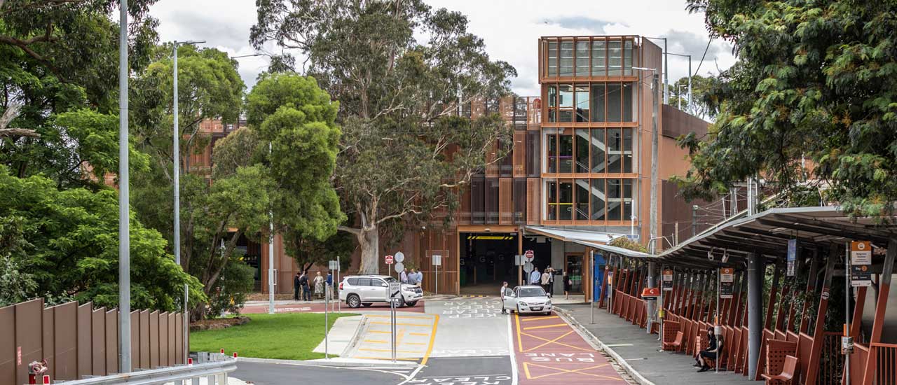 People being dropped off from two cars outside of multi-level  Belgrave Station car park. Another person is sitting at a bus shelter. 