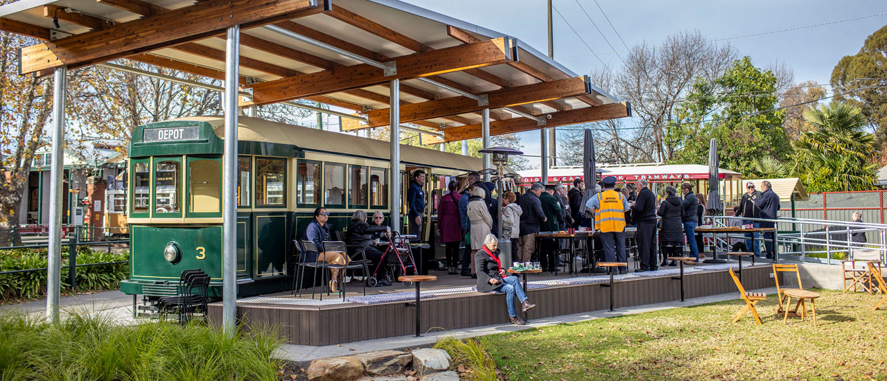 Historic tram has been converted to a cafe at Bendigo Tramways. People can be seen sitting at tables in front cafe while other queue to purchase items. The tram is green and sits under a canopy.  