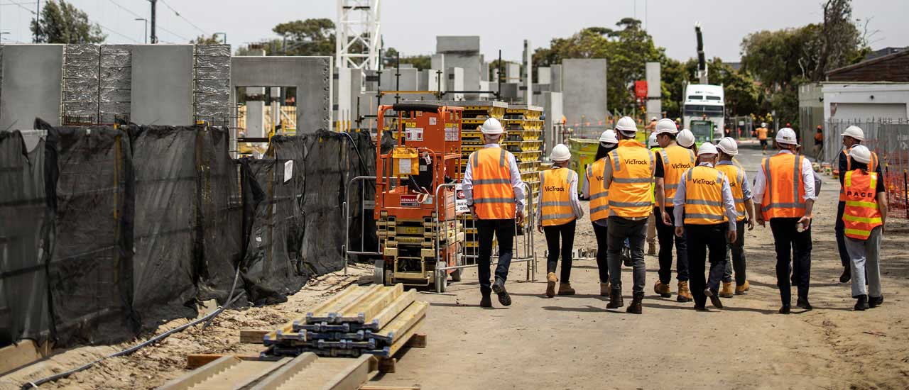 Group of visitors to a construction site walking into the distance wearing high vis and white hard hats.