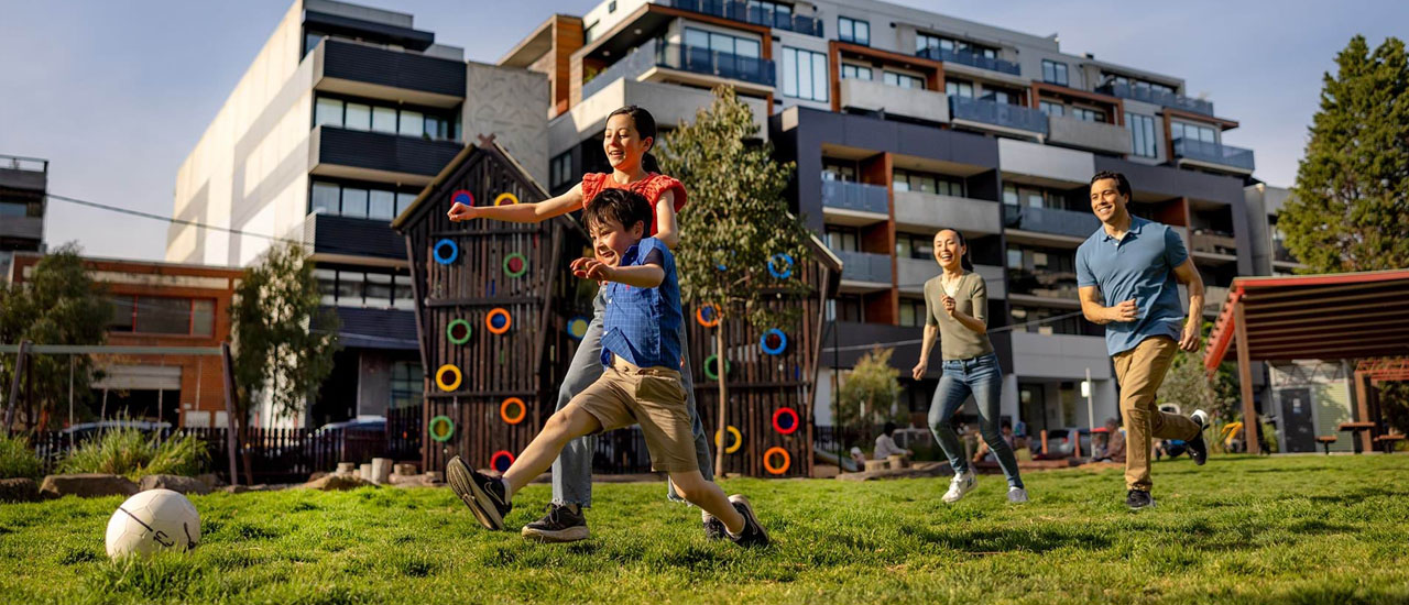 A family of four (two parents and two children) play soccer in a park next door to some medium high rise apartments. There is a wooden playground in the park.