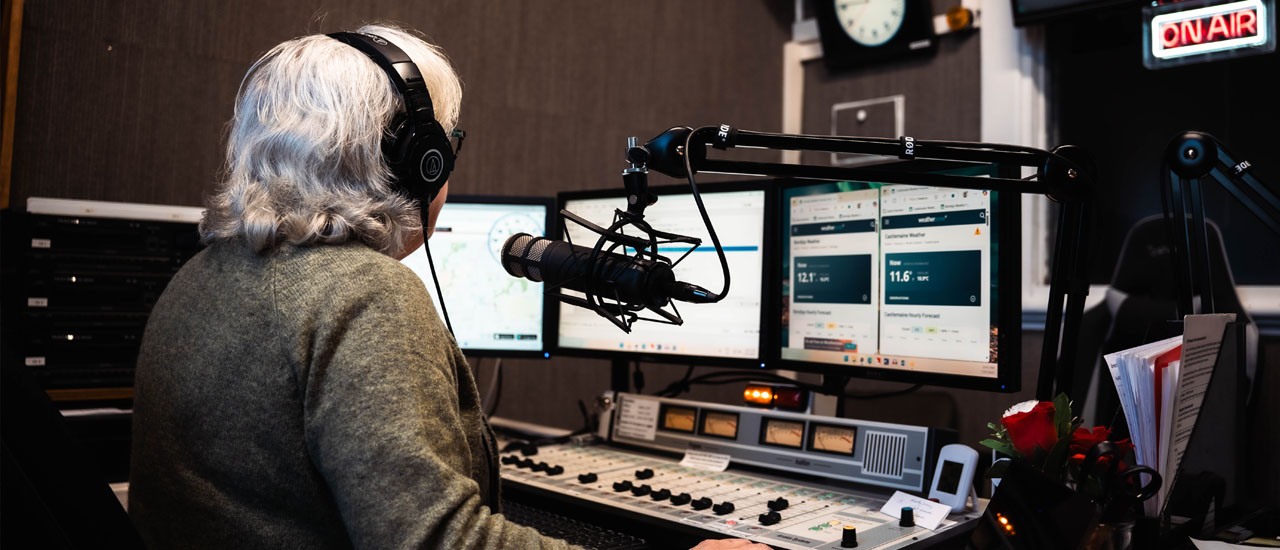 A radio presenter wearing headphones sits at the control board in the radio studio. Computer monitors and microphone can be seen.