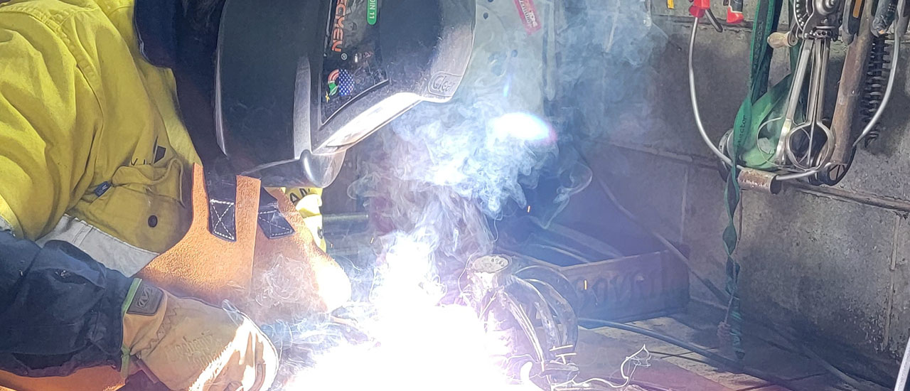 Image of a person using welding tools inside a workshop. The person is wearing a safety mask and high-vis yellow jacket and gloves.. 