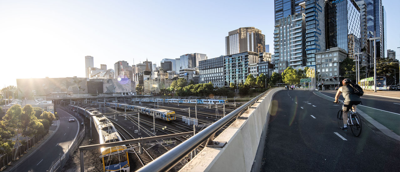 A metro train runs around a bend in front of the Melbourne skyline. A cyclist rides a bicycle across an overpass above the train. A second train, buildings and blue sky can be seen in the background. 