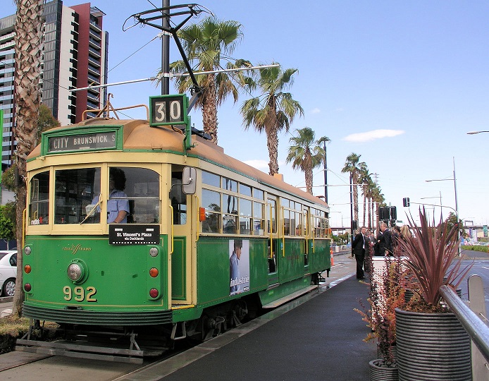 Green W Class tram 992 at a tram stop on route 30 in Melbourne. Blue sky and palm trees are in the background. 