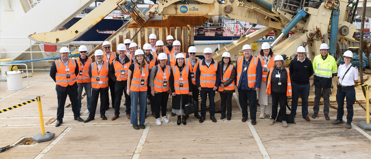 A group of people dressed in orange high visibility vests and white hard hats are looking towards the camera. Construction equipment can be seen in the background. 