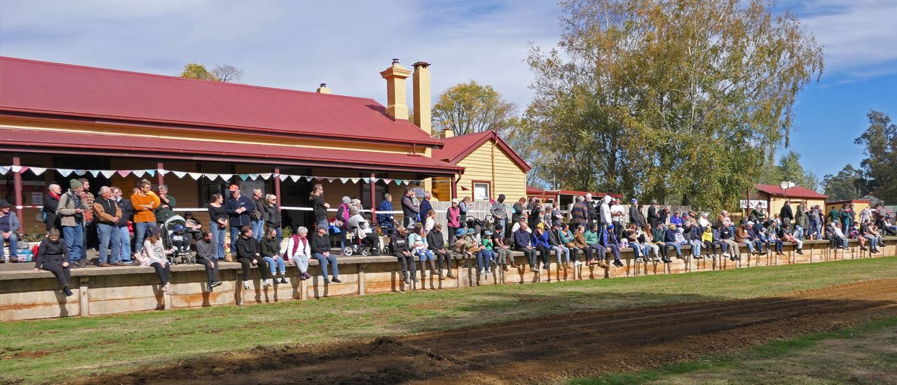 A large group of people sit or stand on the platform of the historic railway station in Trentham Victoria. The yellow timber building has a red tin roof. 