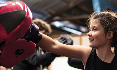 A young child practising boxing in a gym. Child wearing boxing gloves and punching trainer's mits. 
