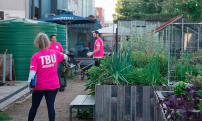 Three people wearing pink TShirts work in an urban garden setting surrounded by raised garden beds with green plants.