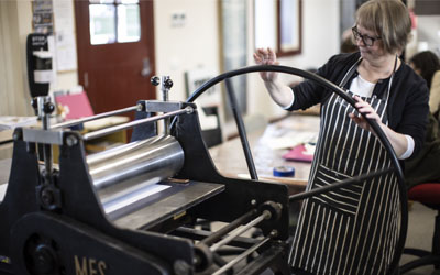 A person wearing a black and white striped apron turns the wheel on a small printing press to produce a print.