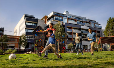 A family of four (two parents and two children) play soccer in a park next door to some medium high rise apartments. There is a wooden playground in the park.