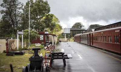 Red heritage railway carriages alongside Moorooduc railway station platform. Cloudy sky in the background.