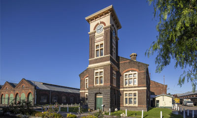 Image of the historic Newport Clocktower building. Railway sheds can be seen next to the building. Blue sky and a tree in the background. 
