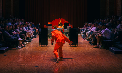 A dancer wearing a red costume performs on a stage in front of an audience. A DJ and music equipment including large speakers can be seen. 